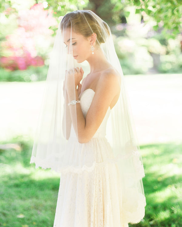A bride in a sunlit field, wearing a two-layer lace veil with blusher. Fingertip length.