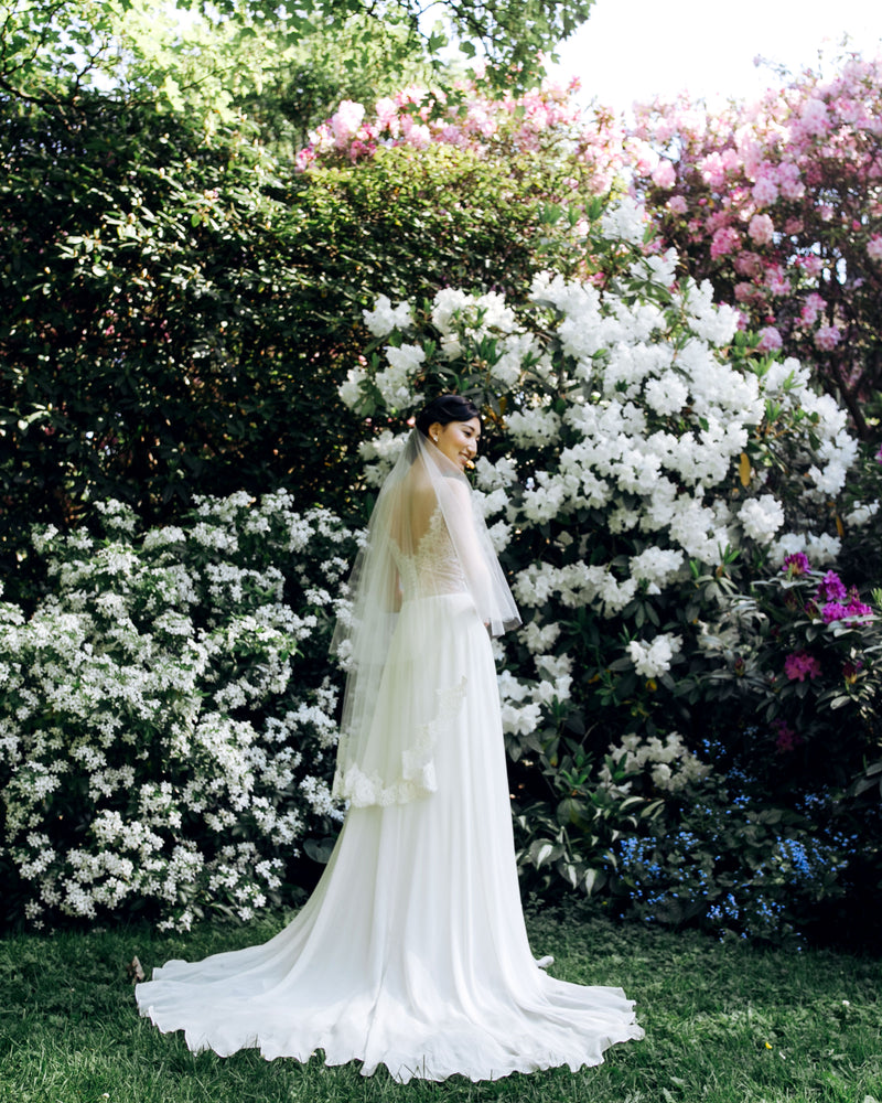 A model stands in a front of blooming hedges, wearing the Azalea lace bridal veil.
