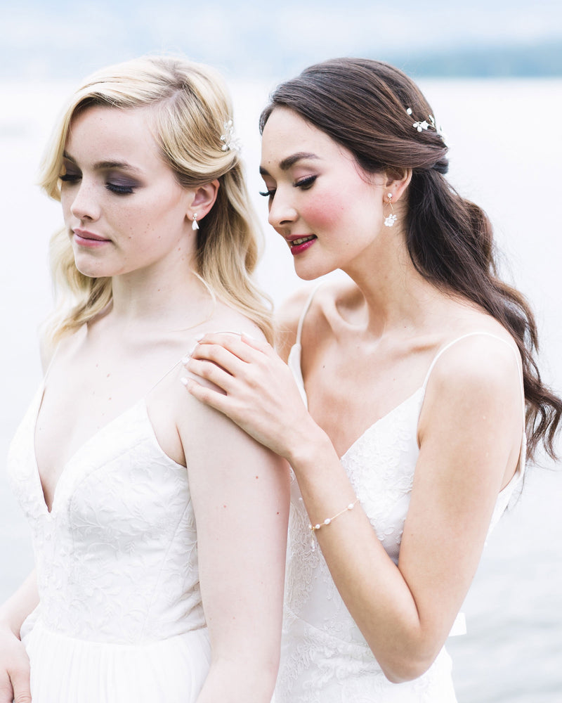 Two models posed together, both wearing bridal accessories with flower details. On the left, the model wears tiny drop earrings with white clay flowers and pearls. On the right, model wears delicate statement earrings and a dainty pearl bracelet.