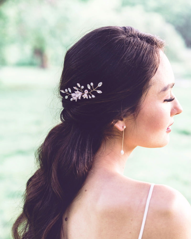 A side view of a model with long dark hair wearing a hair pin in the side of her half up hairstyle. She is also wearing long earrings with pearls at the bottom of a delicate chain.