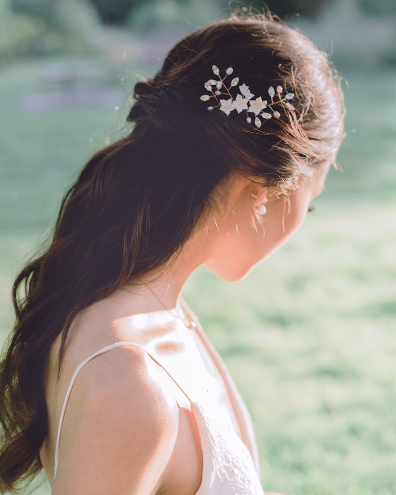 A model with long dark hair poses at golden hour. She is looking down and showing the trio of hair pins styled into her half-up hairstyle. The hair pins are rose gold, with blush flowers, pearls, and crystals.
