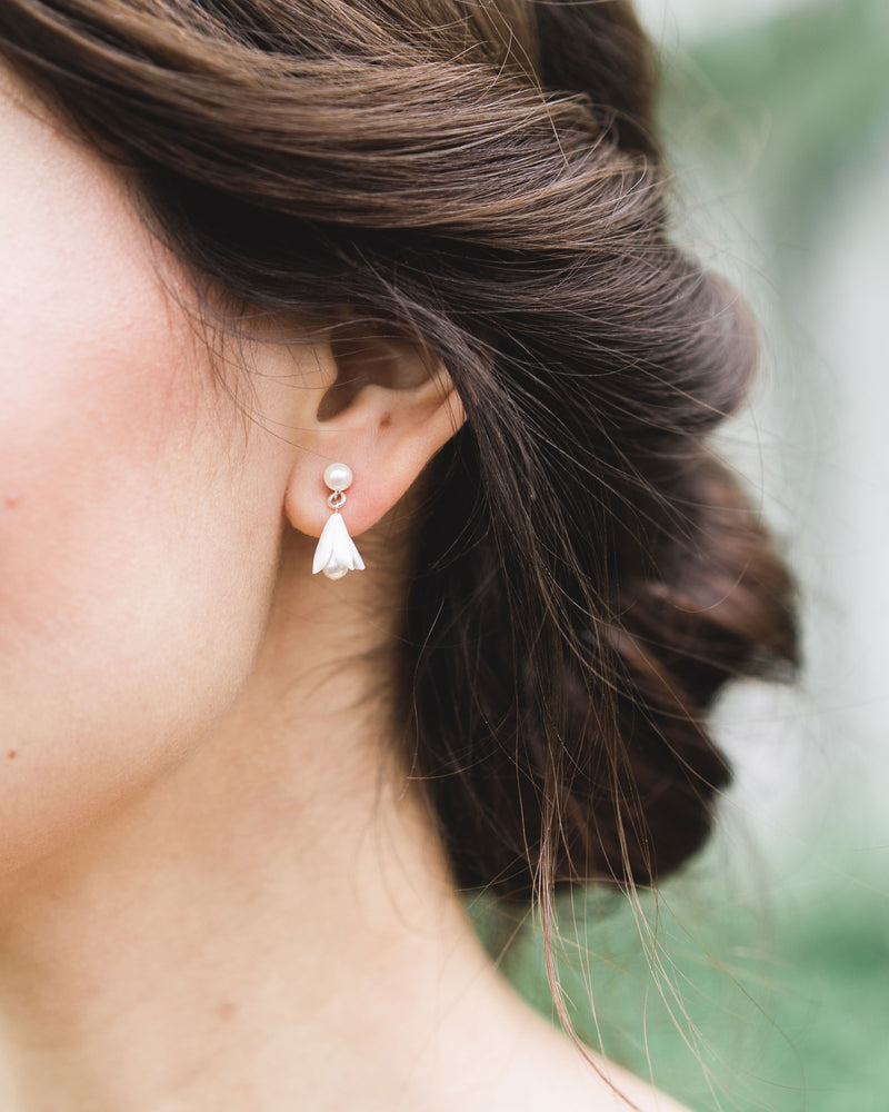 Close up of a dark-haired model with low bun updo and drop earrings; she is wearing the Belle Fleur Petite Earrings, which feature a small flower drop beneath a pearl stud.
