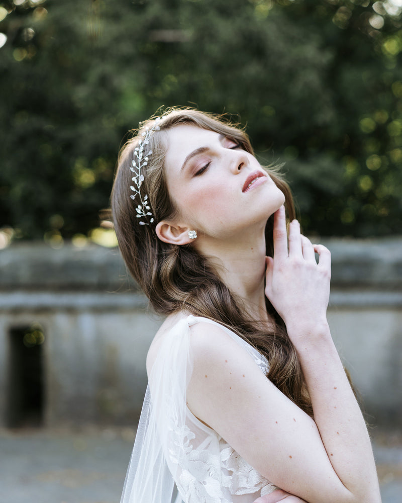 A model poses gracefully with a hair vine in her auburn hair. She wears crystal stud earrings and a tulle bridal cape.
