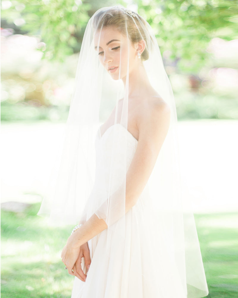 A bride poses in a sunlit field. The blusher of her two-layer veil is worn to the front. She is wearing the Delphine Veil in fingertip length.