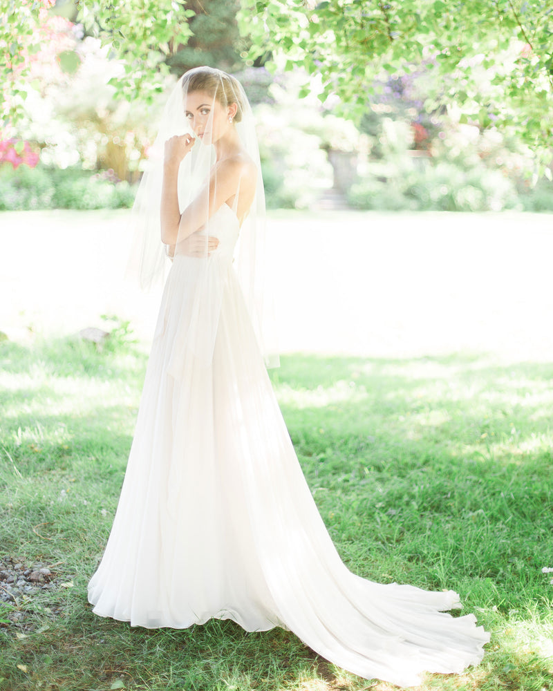 A bride poses in a sunlit field. The blusher of her two-layer veil is worn to the front. She is wearing the Delphine Veil in fingertip length.