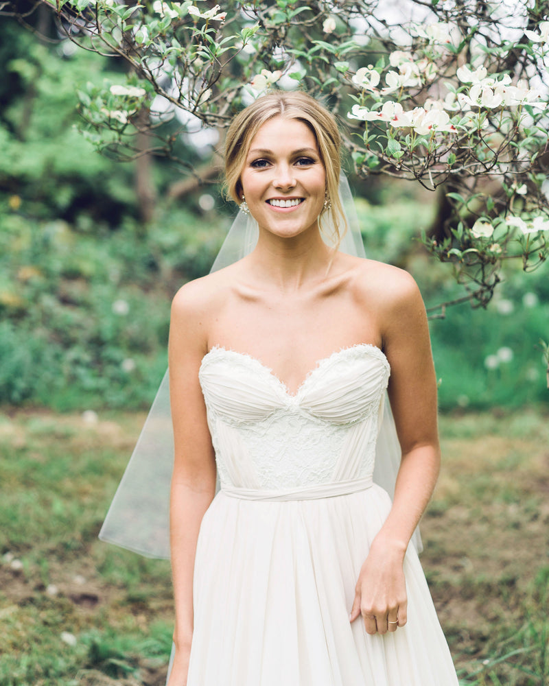 A bride smiles into the camera. Our Delphine Gathered Veil is styled into her romantic bridal updo.