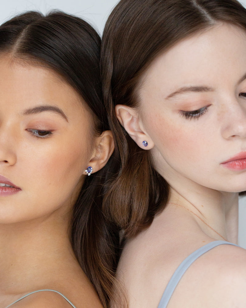 Two models wear bridesmaid jewelry. On left, she wears the Celestial Crystal Stud Earring in rose gold/tanzanite. On the right, she wears the Starry Eyed Stud Earrings in tanzanite.