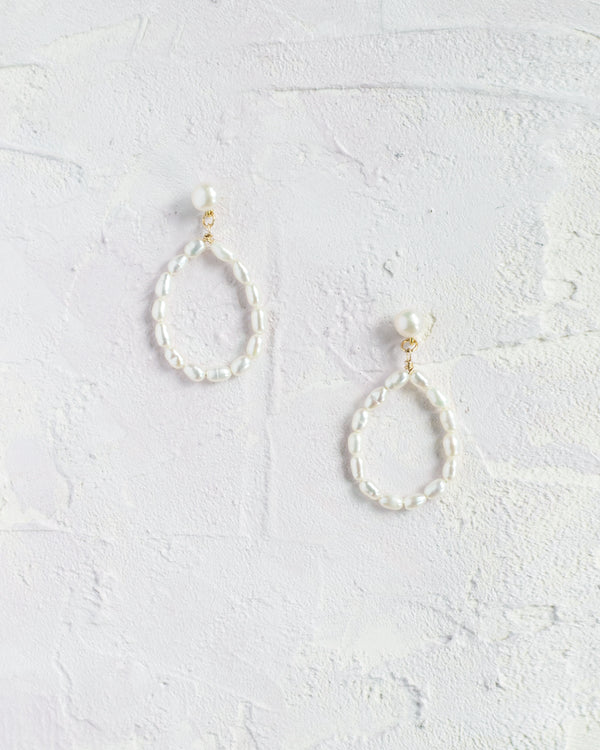 Flatlay view of the Mire pearl earrings in gold. Teardrop shape earrings made of pearls, hanging from a pearl stud.