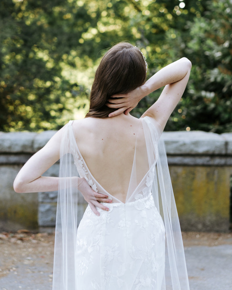 A close back view of a model wearing the Aster Cape Veil. The veil attaches to the straps of her low V-back.