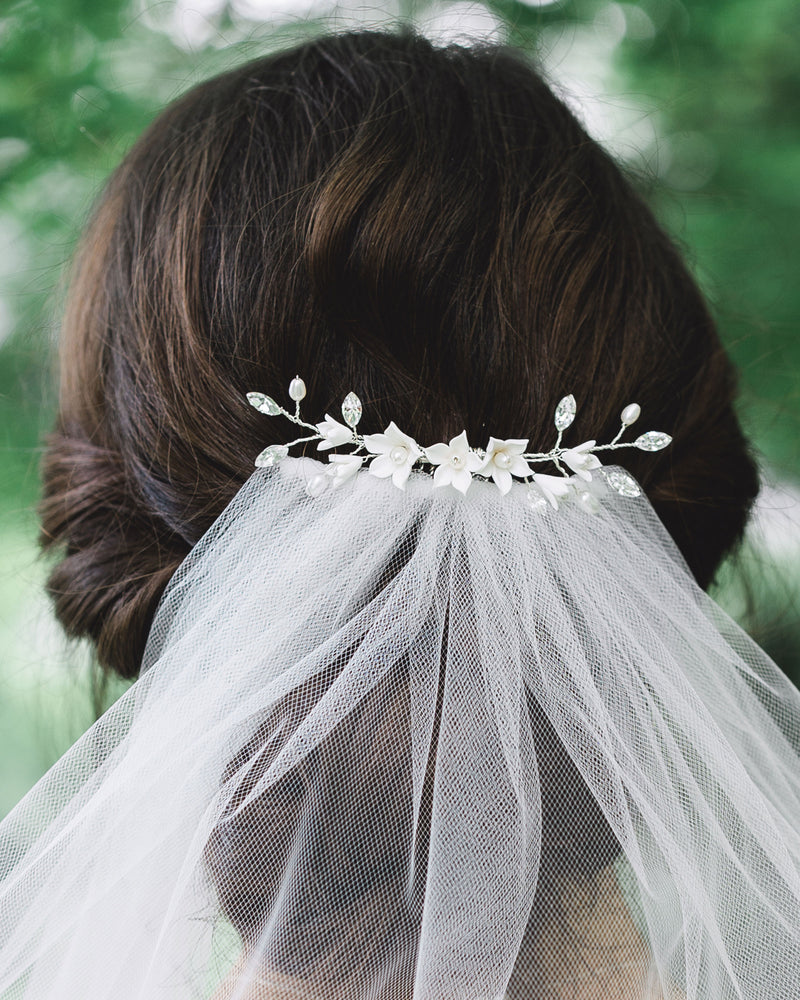 Close-up of model with dark hair and low updo wearing a bridal hair comb with crystals, white flowers, and pearls over a simple veil.