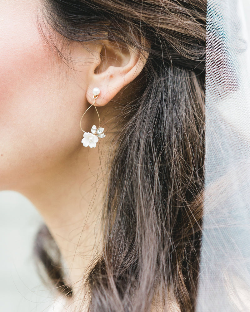 Close up of model wearing delicate statement earring with white flower, crystals, on a wire teardrop hanging from a pearl stud.