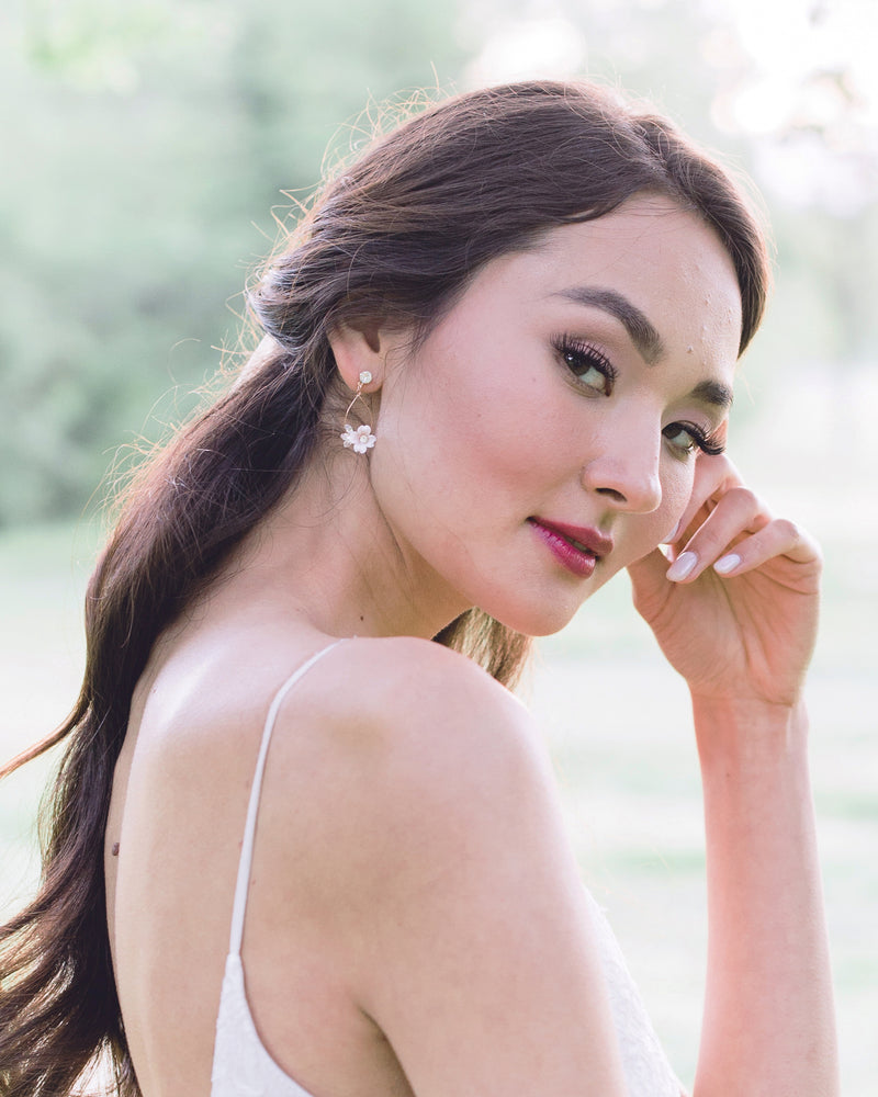 Model with dark hair and a dark red lip is smiling softly and modelling the Belle Fleur Earrings with rose gold wire, blush flowers, and crystal accents.