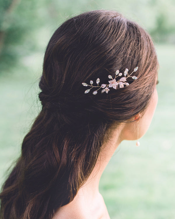 A back view of a model with dark hair and a half-up hairstyle. She is wearing a hair pin in the side that is gold with blush flowers, pearls and crystals.