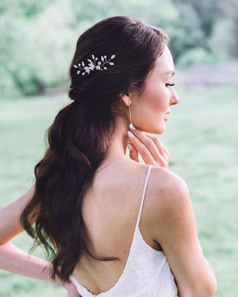 A side view of a model with long dark hair wearing a hair pin in the side of her half up hairstyle. She is also wearing long earrings with pearls at the bottom of a delicate chain.