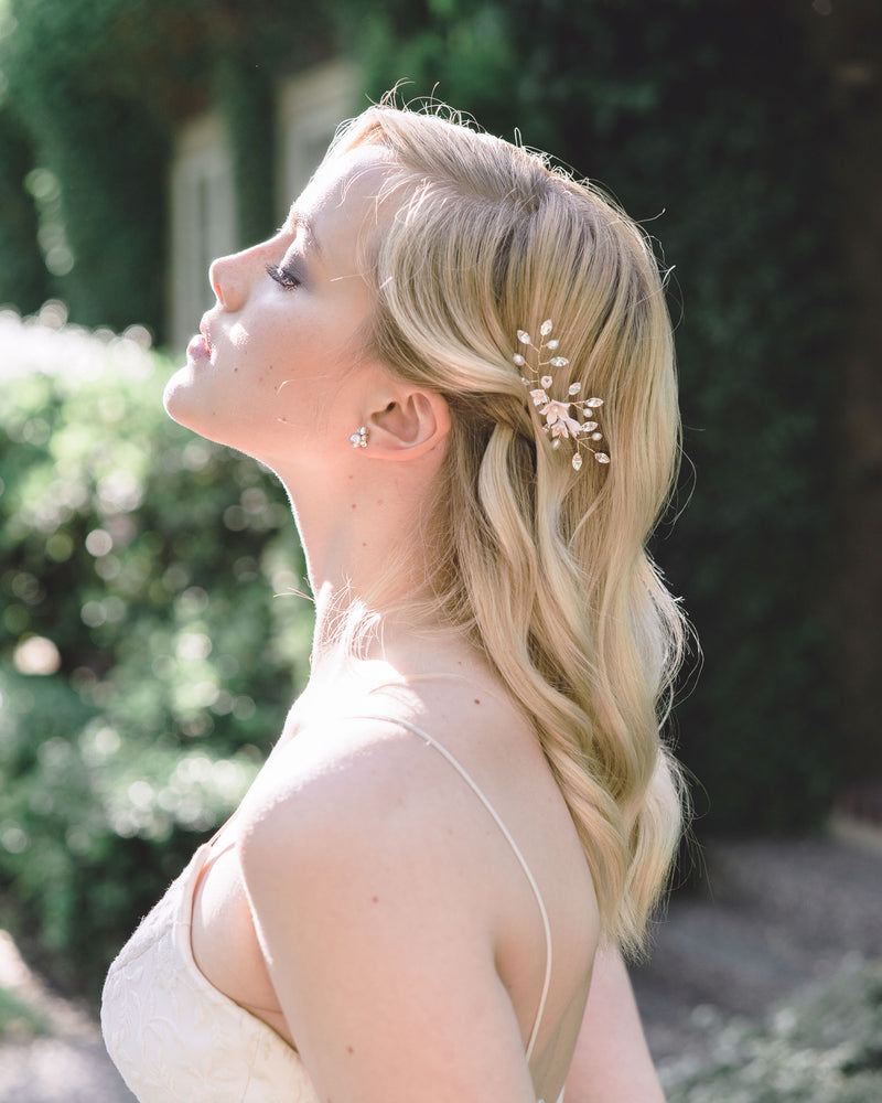 A model raises her face to the sun. She has blonde hair styled in soft waves and a hair pin styled above the ear. The hair pin is gold, with blush flowers and crystals. She is wearing stud earrings with a cluster of crystals above a blush pearl.