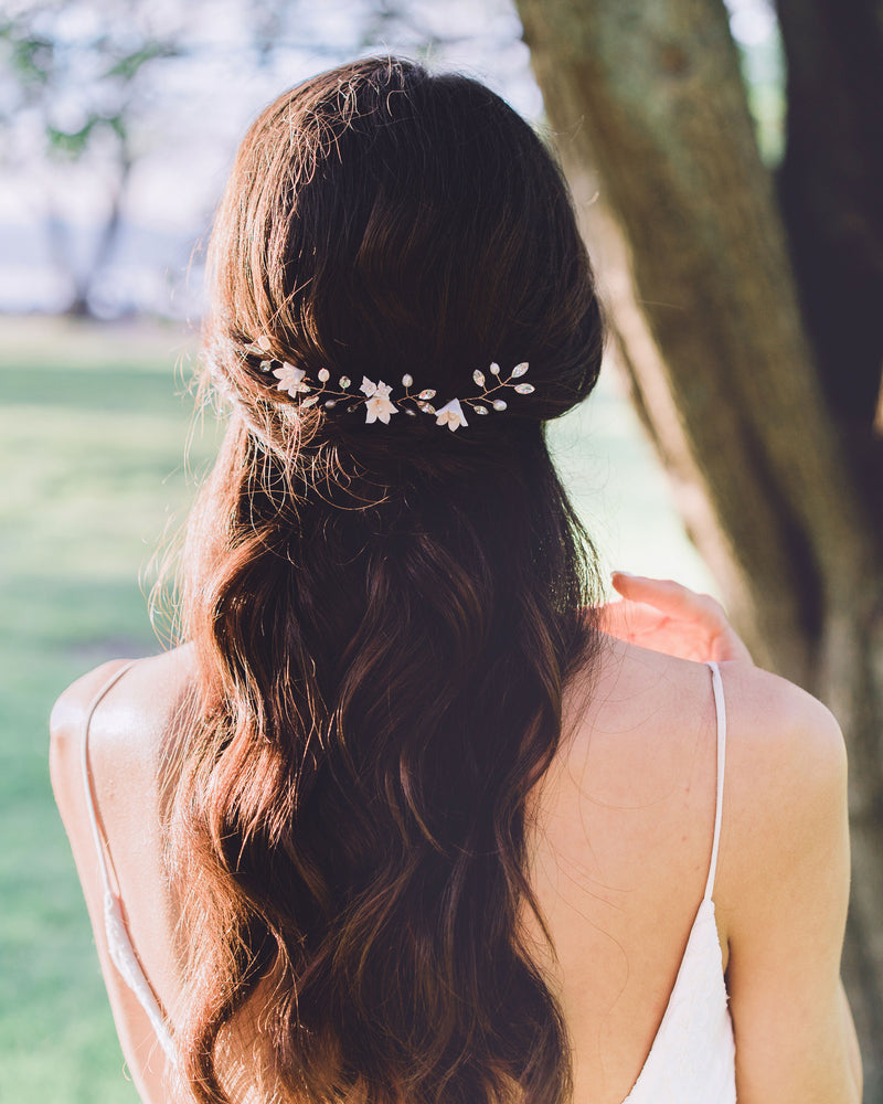 A back view of a model with long dark hair wearing a trio of hair pins in her half up hair. The hair pins have been placed along the back of her updo, so that it styles across the back of her hair. The hair pins are rose gold, with blush flowers, pearls, and crystals.