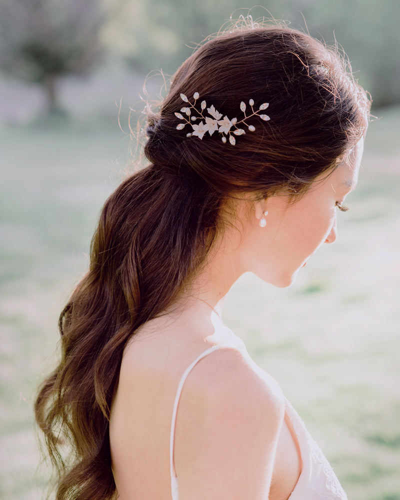 A model with long dark hair styled into a half updo is standing to the side and looking down gracefully. She is wearing a trio of hair pins in her hair and pearl drop bridal earrings.
