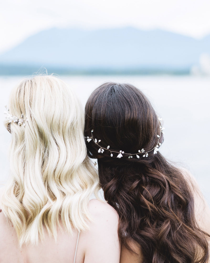 Two models face the ocean with their backs to the camera. The dark haired model on the right wears a dainty hair vine styled across the back of her half-up hairstyle. The hair vine has flowers, pearls and crystals.