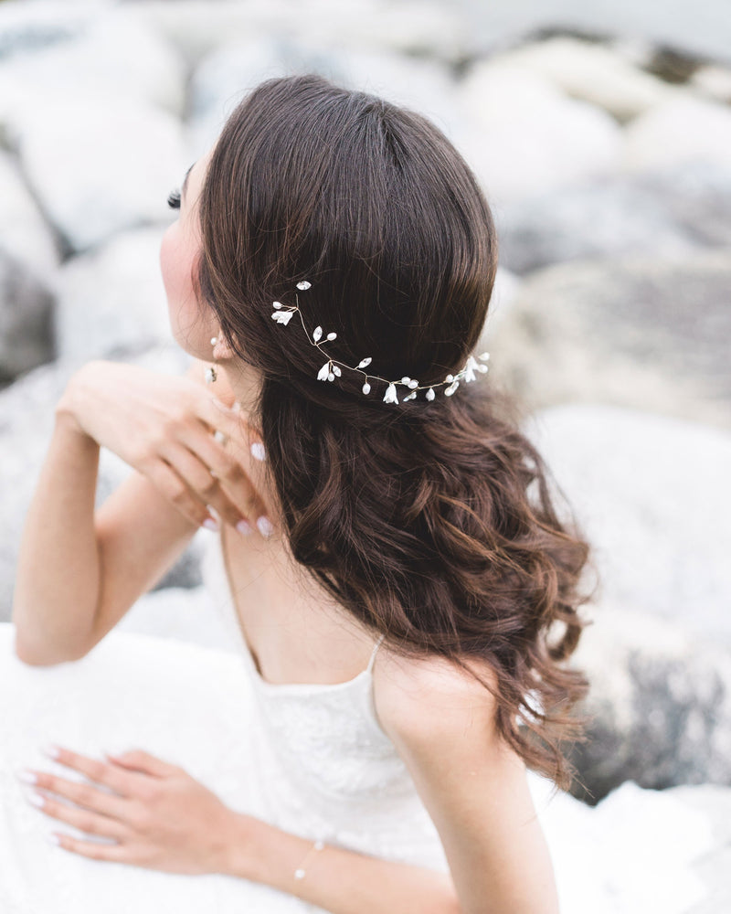A model poses on the rocky shore. She is wearing a wedding dress and showcasing a hair vine styled in the back of her half-up hairstyle. The hair vine has flowers, pearls, and crystals.