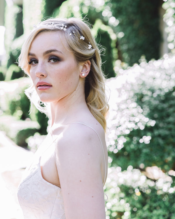 A blonde model with smoky eyes gazes directly into the camera. She is wearing a delicate hair vine made of flowers, pearls and crystals. She is also wearing classic pearl studs.