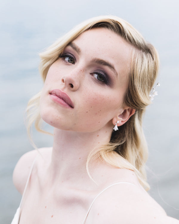 A model with blonde hair and smoky eye makeup gazes into the camera. She is wearing the Petite Belle Fleur Earrings, which have a tiny flower drop falling from a freshwater pearl stud.