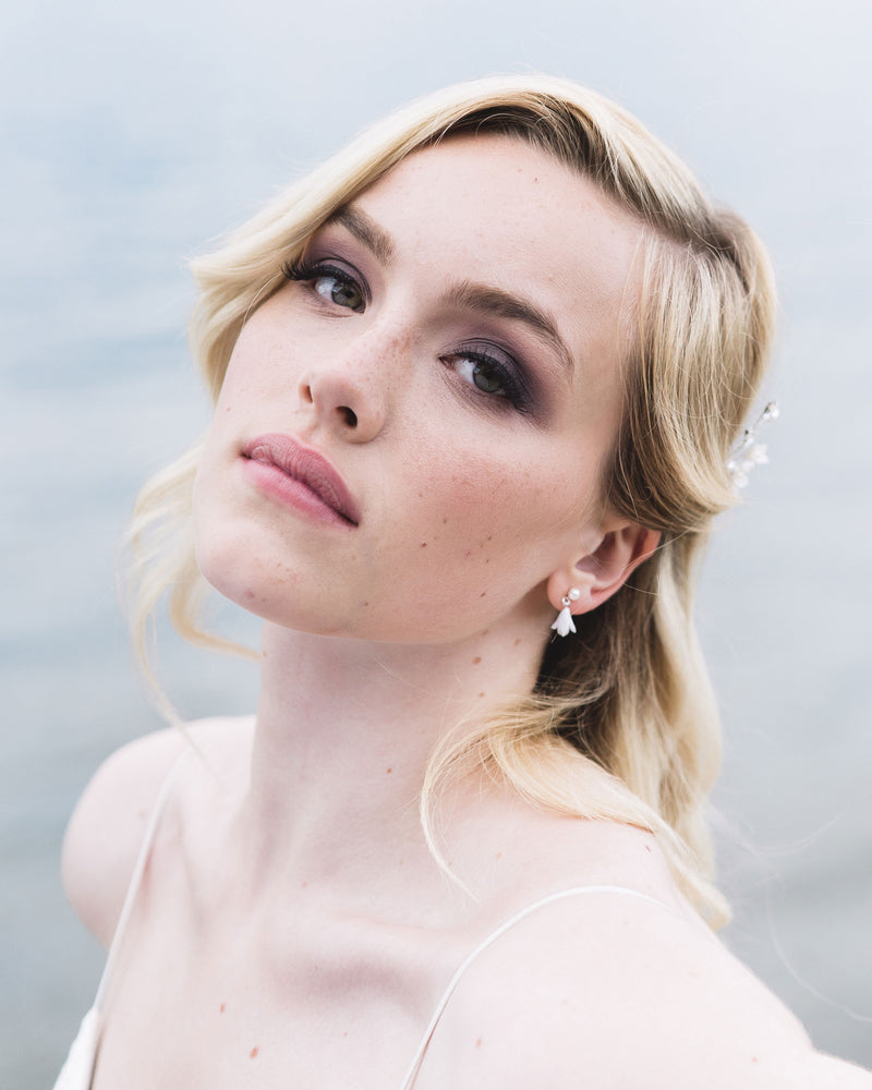 A model with blonde hair and smoky eye makeup gazes into the camera. She is wearing the Petite Belle Fleur Earrings, which have a tiny flower drop falling from a freshwater pearl stud.