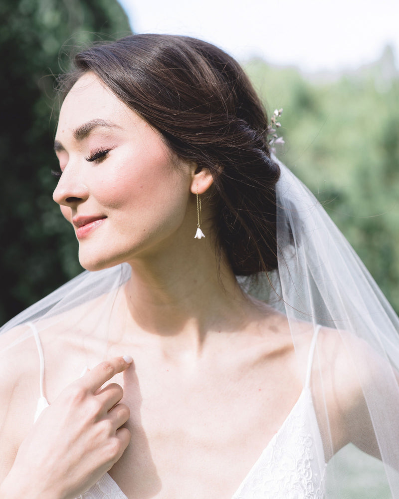A dark-haired model smiles with her eyes closed and turns her face to the sun. She is wearing delicate ear threader earrings with tiny blush flower drops. A veil is styled into her low updo.