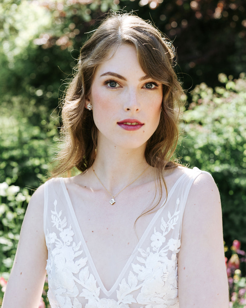 An auburn-haired bride is wearing a delicate crystal jewelry set. She wears crystal stud earrings and a delicate gold necklace with a crystal drop.