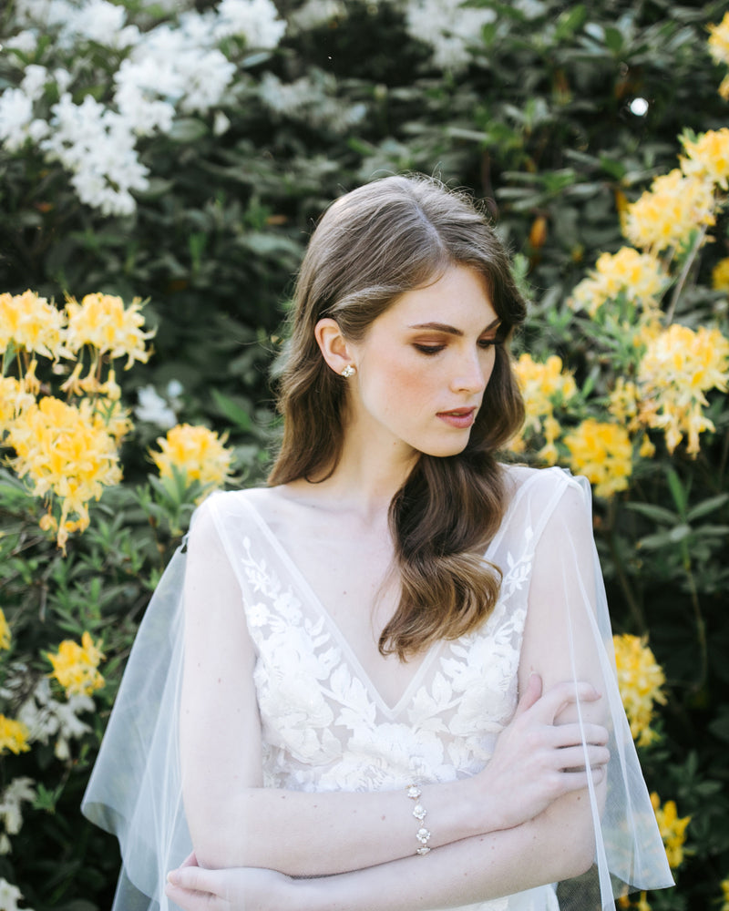 A model poses in a wedding dress and tulle bridal cape, wearing the Celestial Pearl Cluster Bracelet in gold and matching pearl stud earrings.