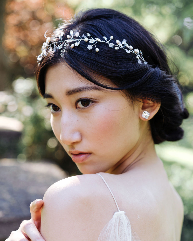 A model gazes into the distance.  She wears crystal stud earrings. Her black hair is styled in a low updo with twists and a silver hair vine with gemstones, pearls, and crystals.