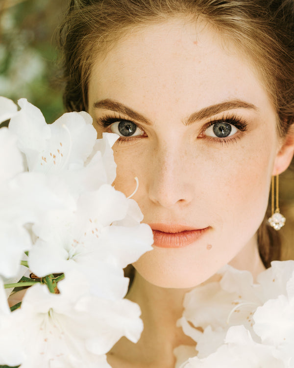A model poses with white flowers all around her. She is wearing the Celestial Threader Earrings in gold with white opal crystals.
