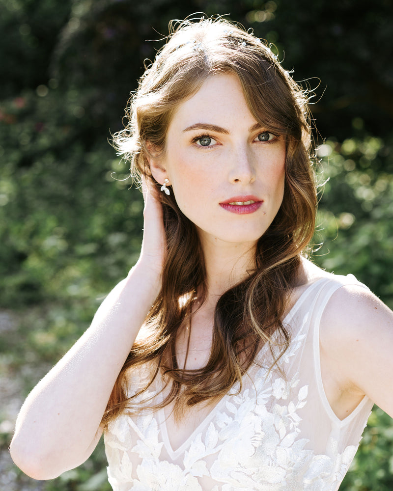 A model with auburn hair stands with sun on her soft bridal waves. She is wearing the crystal leaf earrings in gold.