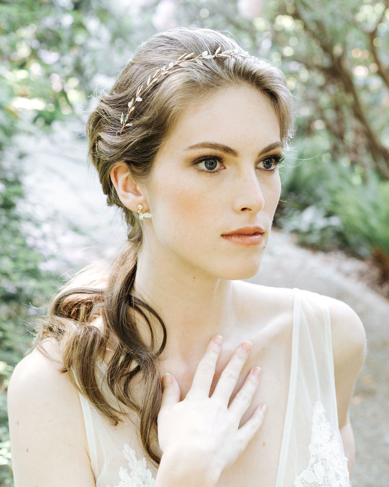 A model stands in a dream-like forest with her hair in a low bridal ponytail. She wears the Crystal Leaf Earrings in gold and the Sea Mist Hair Vine with crystals and gemstones.