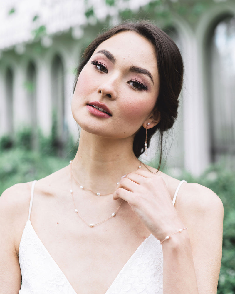 A model is wearing a pearl jewelry set. She is wearing a layered pearl necklace with matching dainty pearl bracelet, along with long drop earrings with a trio of pearls.