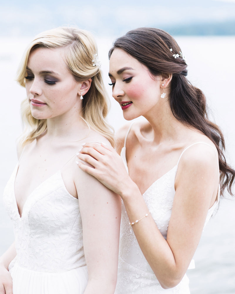 Two models pose along the seashore. They are wearing bridal accessories; the blonde model on the left wears small flower drop earrings and the dark-haired model on the right wears a dainty pearl bracelet and delicate statement earrings.