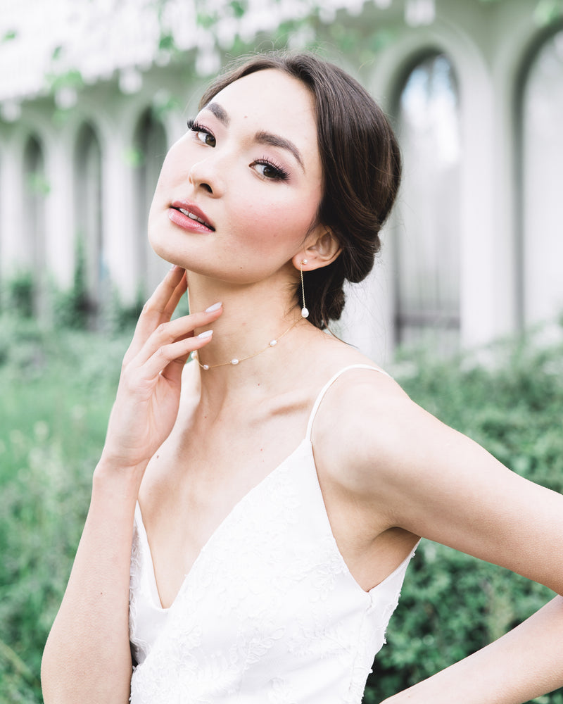 A model with a low bridal updo wears the Dainty Pearl Necklace in gold, paired with the Teardrop Pearl Long Earrings in 2" length.