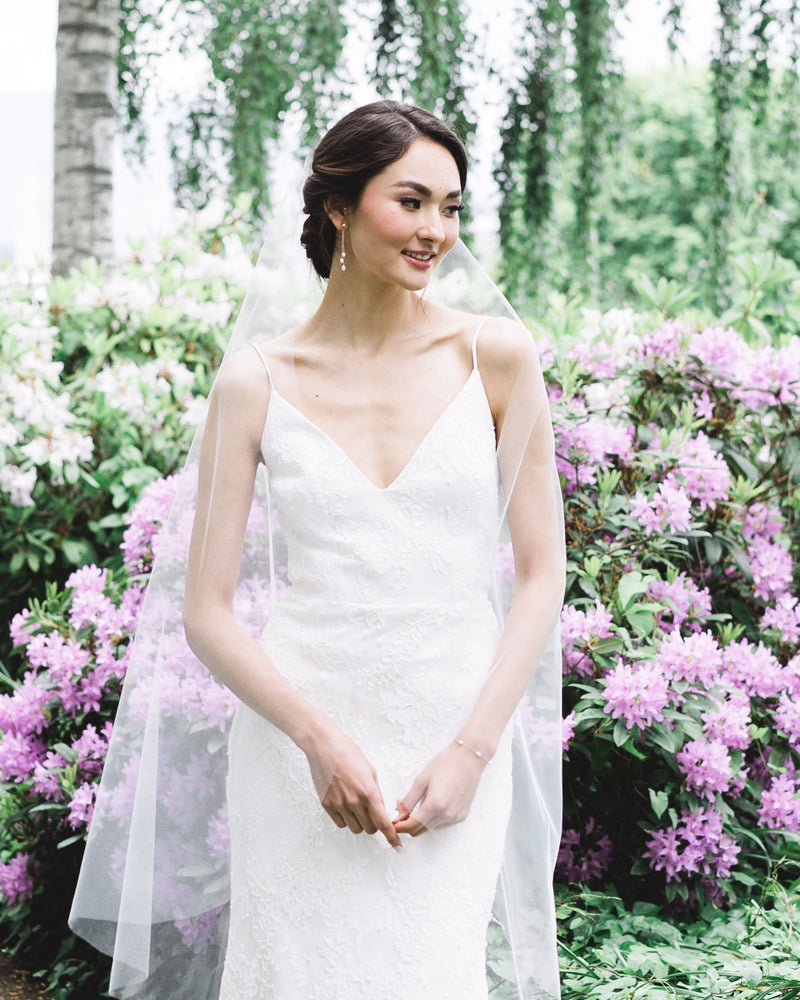 A bride stands in front of blooming shrubs. She wears a soft vel without gathers, the Dainty Pearl Trio Earrings and matching Dainty Pearl Bracelet.