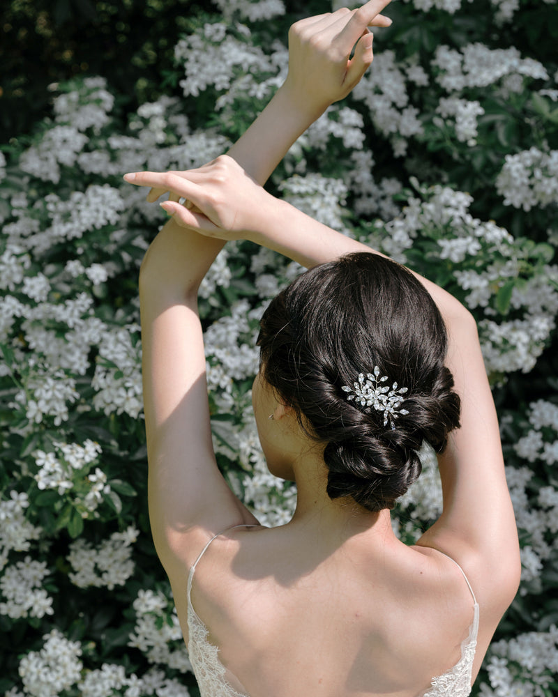 A model poses gracefully in front of a blooming hedge. She is wearing the Delicate Comb in silver and all crystal in her low bridal updo.