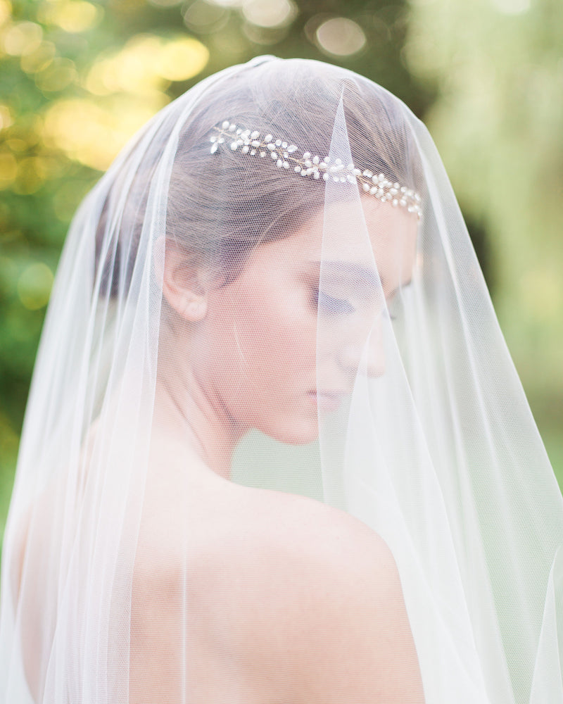 A model is half-turned to the camera. She wears a boho headpiece of pearls and crystals with her bridal updo. A soft veil with the blusher to the front covers her face.