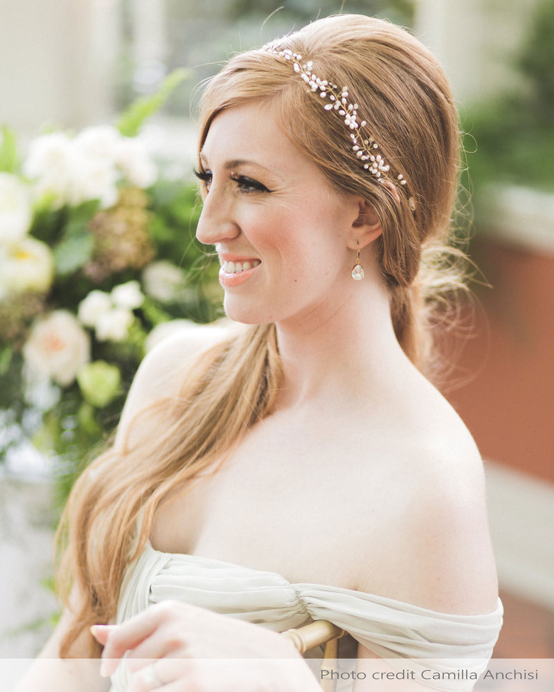 A bride wears her long hair pinned to the side, with a delicate hair vine styled at the front. The hair vine has pearls, crystals, and pearl flowers.