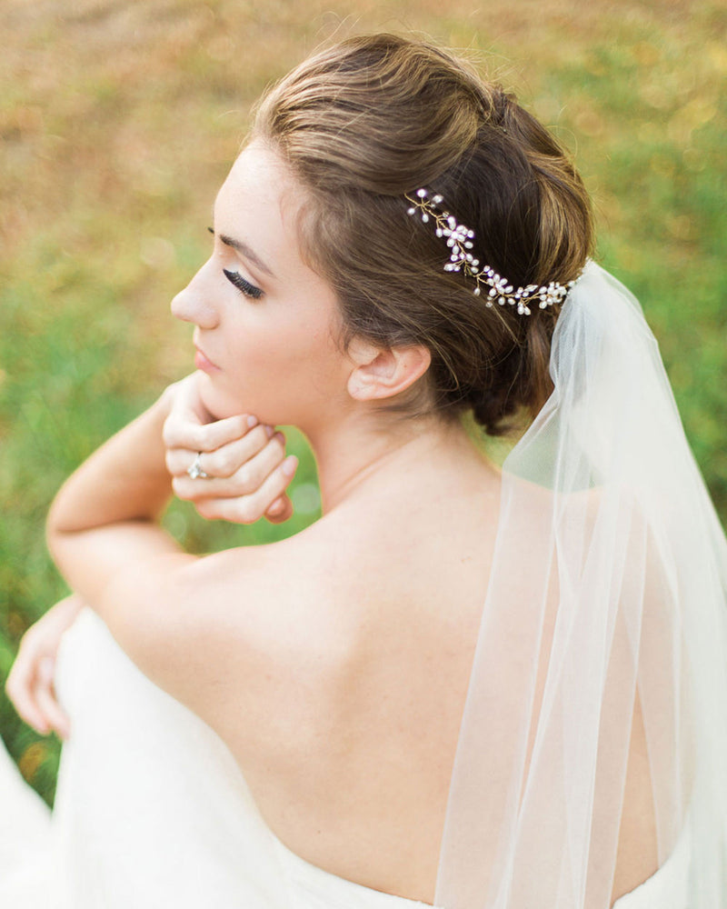 A model sits half turned to the camera. A hair vine in styled into the back of her bridal updo and is paired with a tulle veil. The Delicate Pearl & Crystal Hair Vine has pearls, crystals, and pearl flowers.