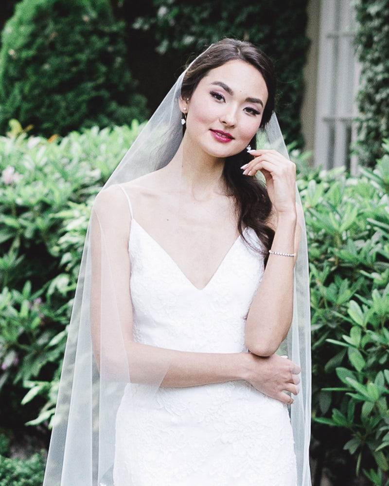 A bride wears our Senna Veil in her half up hairstyle. She wears the Delicate Tennis Bracelet paired with our Belle Fleur Earrings.