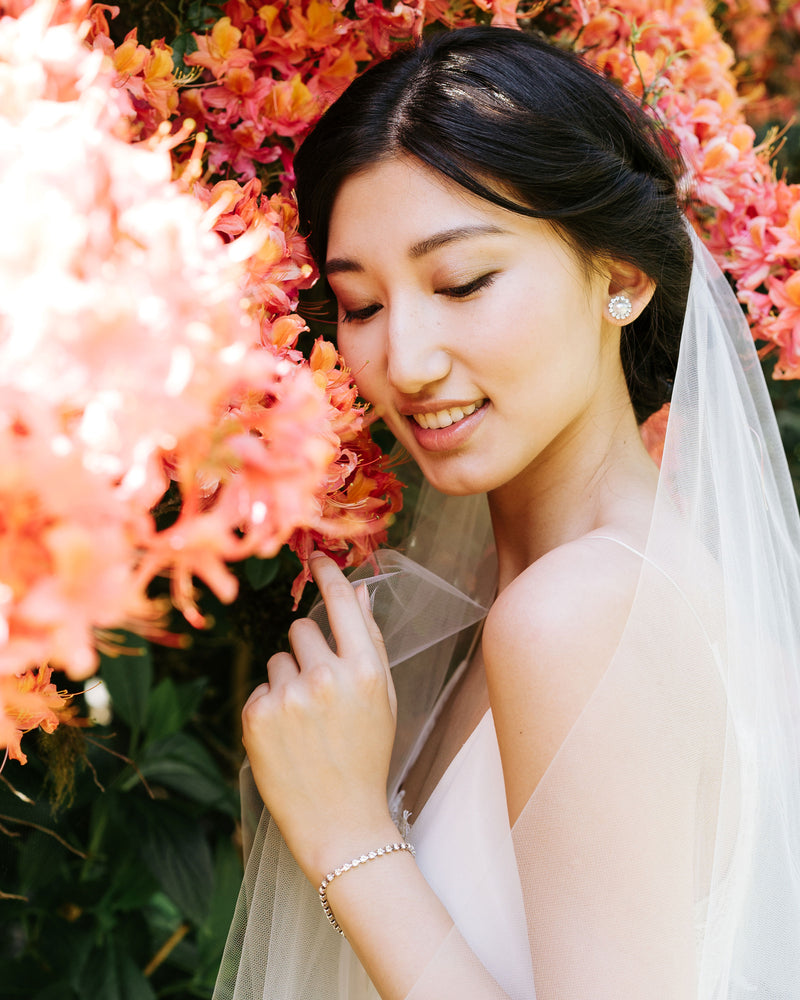 A model poses against a blooming shrub. She wears a soft veil and has a delicate crystal tennis bracelet on her wrist, styled to match the Halo Pearl Stud Earrings she is wearing.
