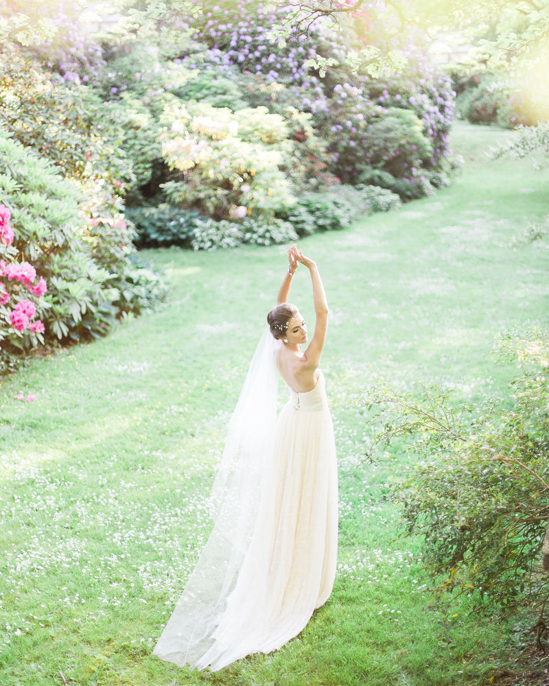A bride dances in a bright, grassy field. Her soft two layer veil trails behind her; she is wearing the Delphine Veil in chapel length.