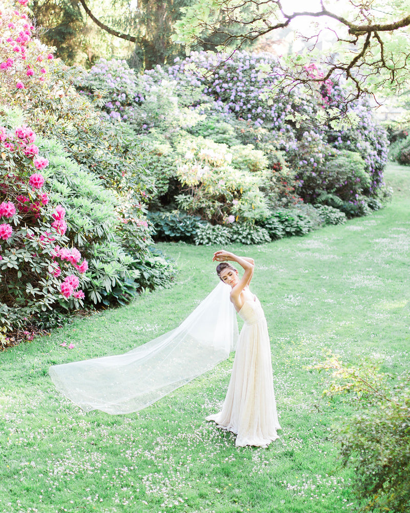 A bride dances in a bright, grassy field. Her soft two layer veil trails behind her; she is wearing the Delphine Veil in chapel length.
