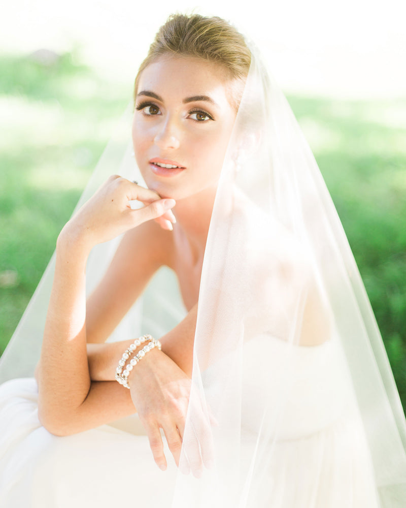 A model sits with sunlight all around. She has a two-layer veil without gathers styled with her bridal updo.