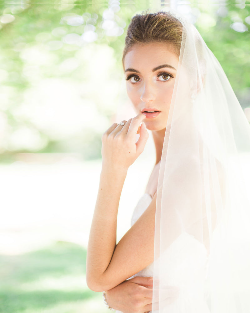 A model poses in a sunlit field. Her two-layer fingertip veil is worn to the back over her bridal updo.