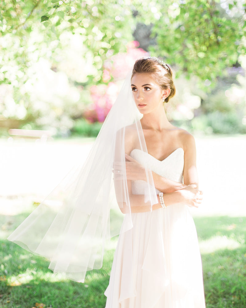 A bride poses in a sunlit field. Her two-layer veil floats around her softly. She is wearing the Delphine Veil in fingertip length.