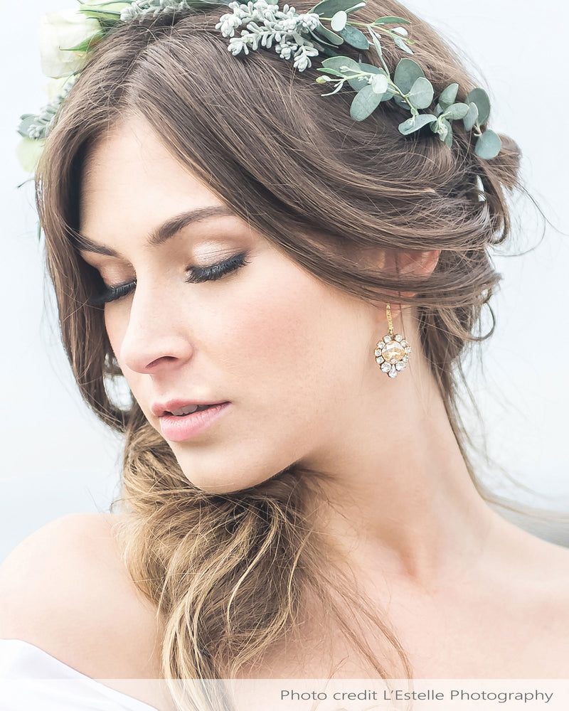A model with a floral crown wears the Enchanted Crystal Drop Earrings in gold with champagne crystal centers. She is wearing the pave crystal hook version.
