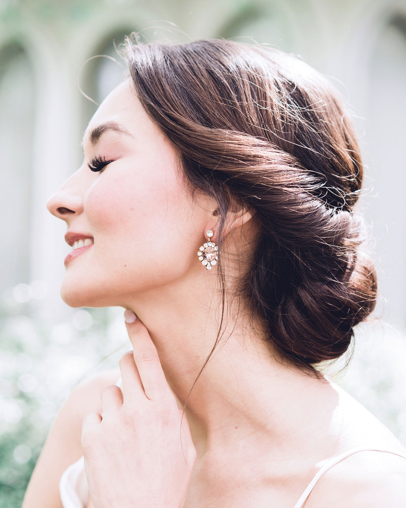 A model smiles as she wears the Enchanted Crystal Drop Earrings in gold with blush crystal centers. Styled with a low bridal updo with twists.